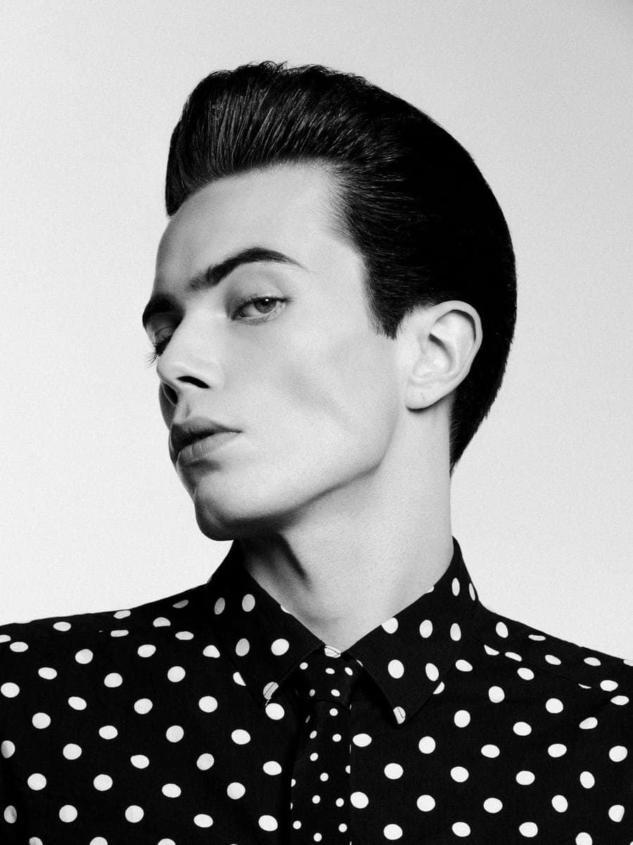 Slicked-back pompadour over a black polka-dot shirt, the sitter turned out of the light