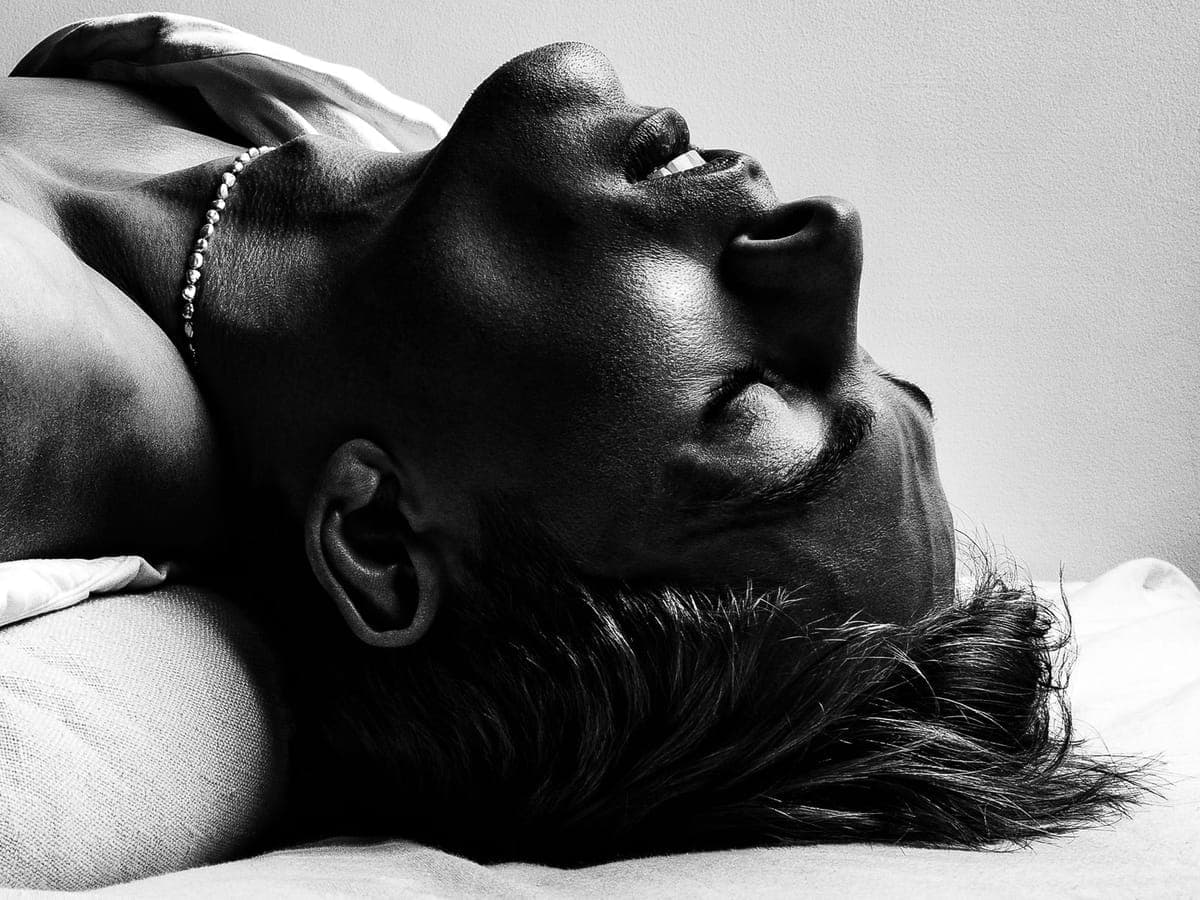Head tipped back in laughter on white linen, a beaded chain at the throat