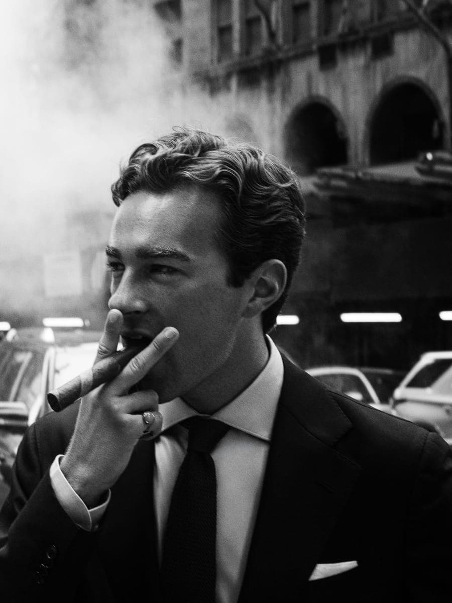 Man in a dark suit drawing on a cigarette, steam and parked cars along the street behind him