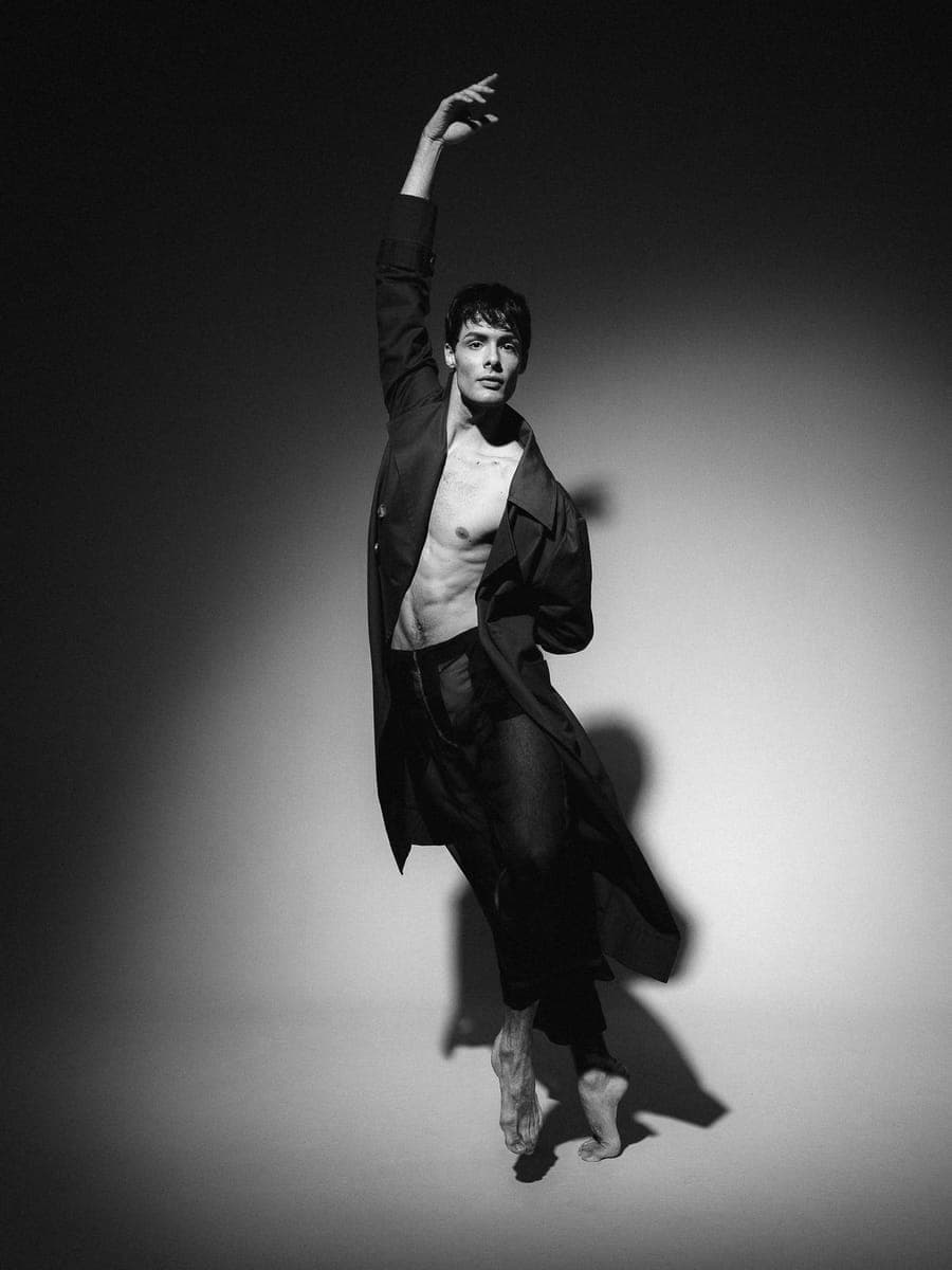 Dancer rising on half-pointe in an open coat and sheer trousers, one arm overhead