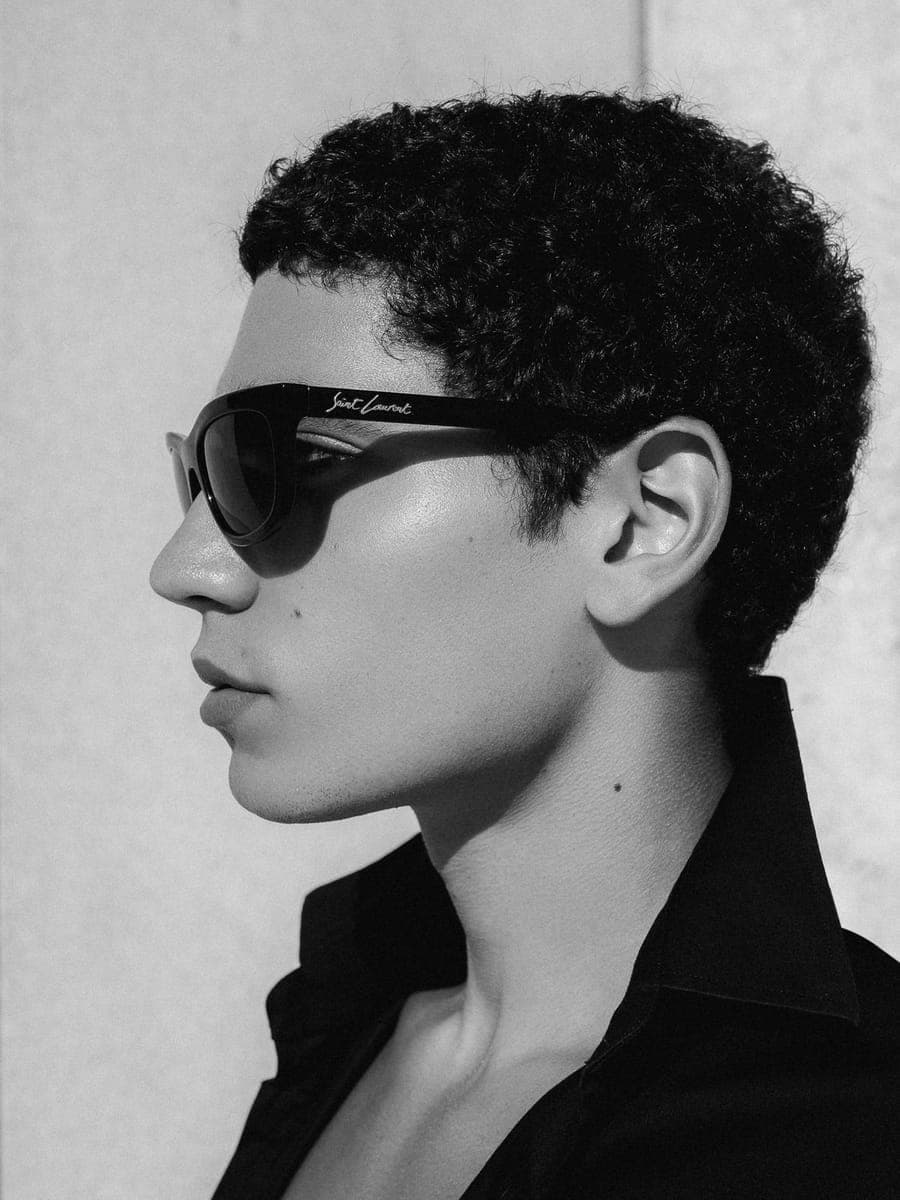 Profile in Saint Laurent sunglasses and an open black collar, lit by hard sun