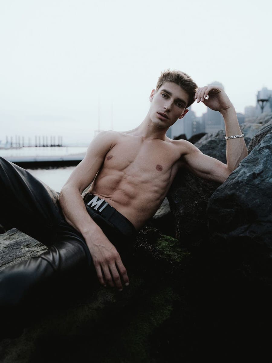 Model in leather trousers reclining on harbor rocks, Lower Manhattan across the water