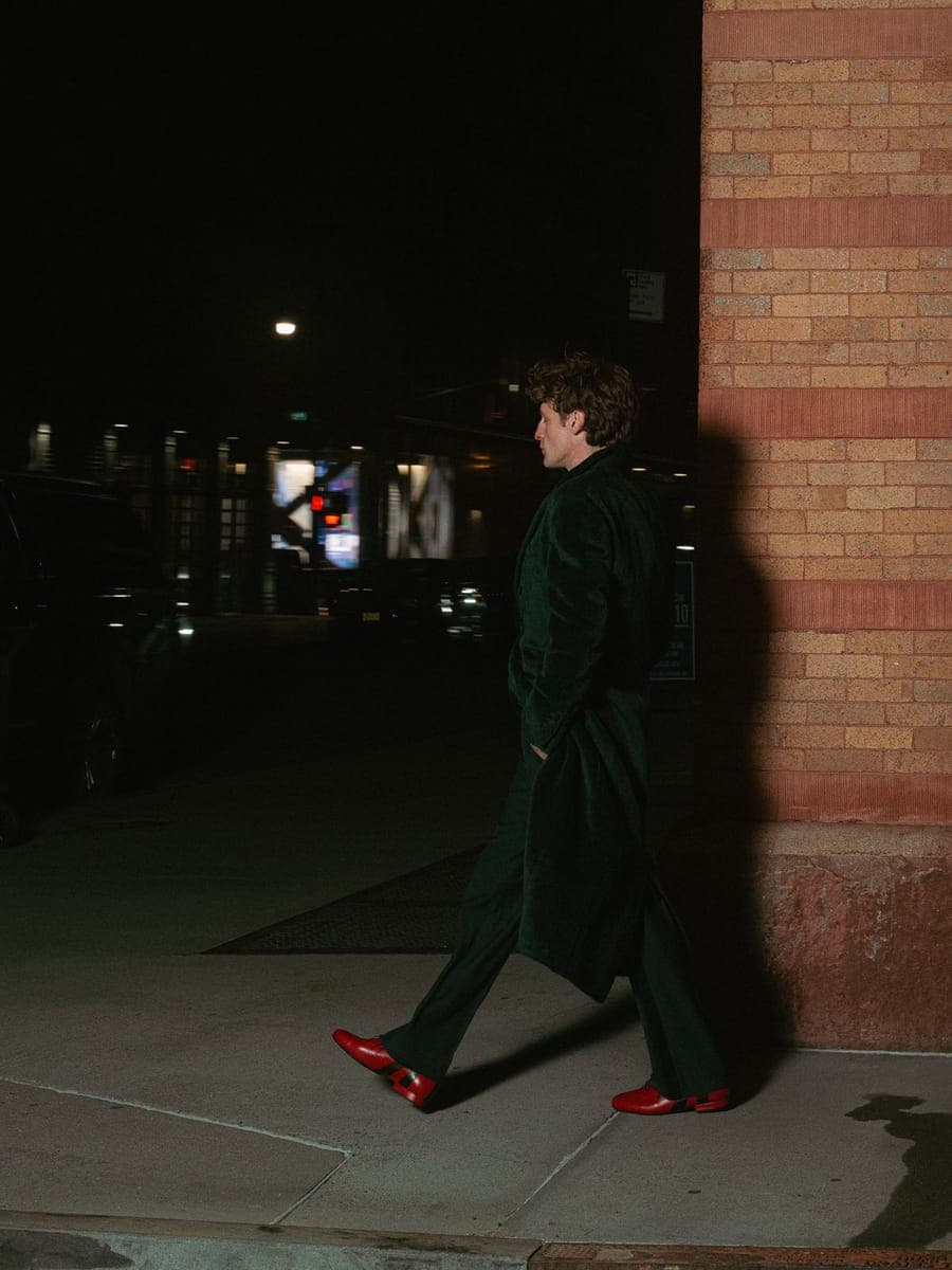 Man in a long green coat and red shoes striding past a brick corner at night